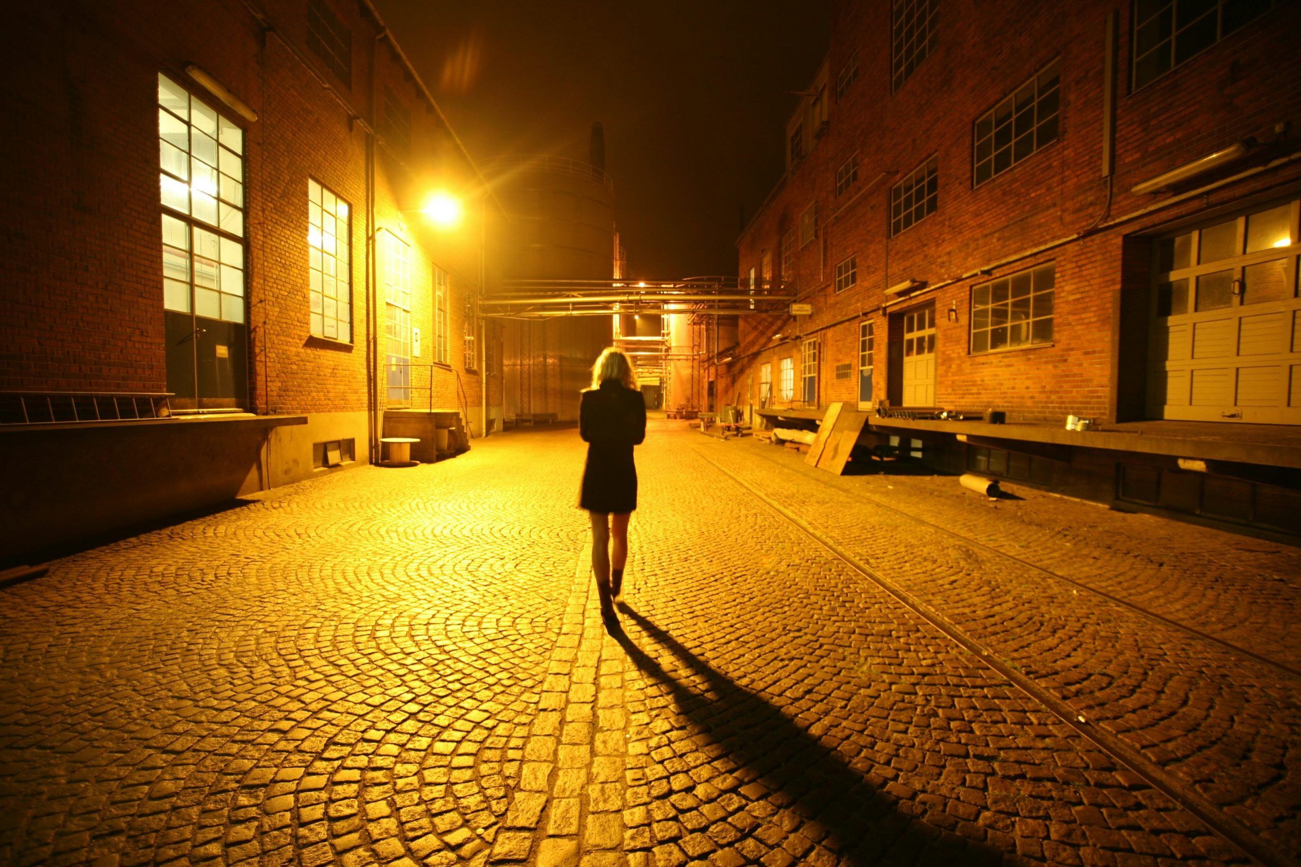 Woman walking alone at night in poorly life and secluded warehouse area, feeling secure because she knows the Life Backup App will check on her and summon help if she doesn't respond.